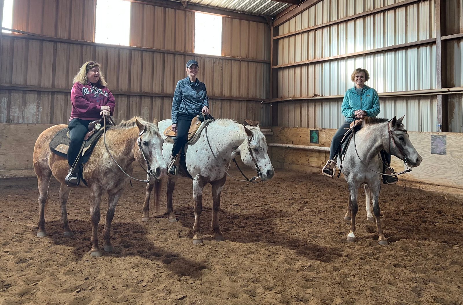 Three adult riders sit on Appaloosa horses in the indoor arena, smiling at the camera.