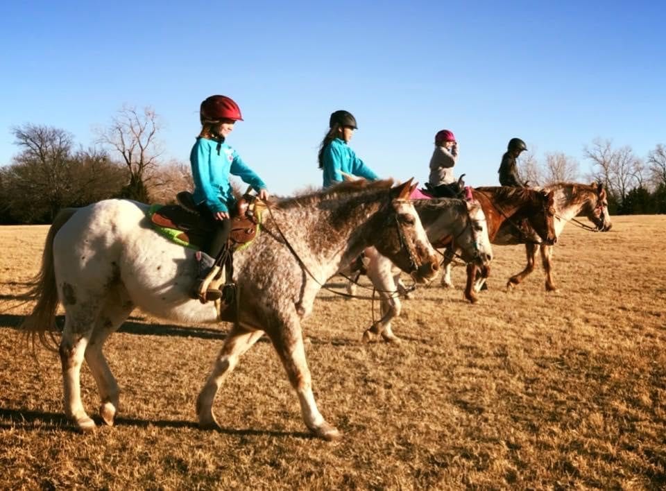 Three young riders in helmets ride side by side through a pasture during a group lesson.