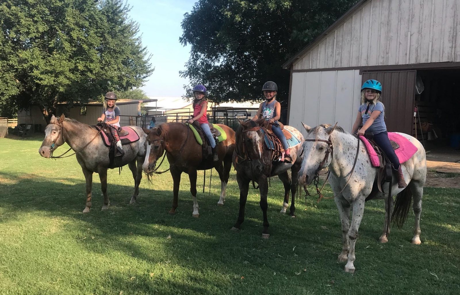 A line of horses and riders heads out on a trail ride past the barn.