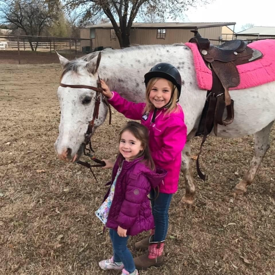Two girls in pink jackets pose with a saddled Appaloosa before a lesson.