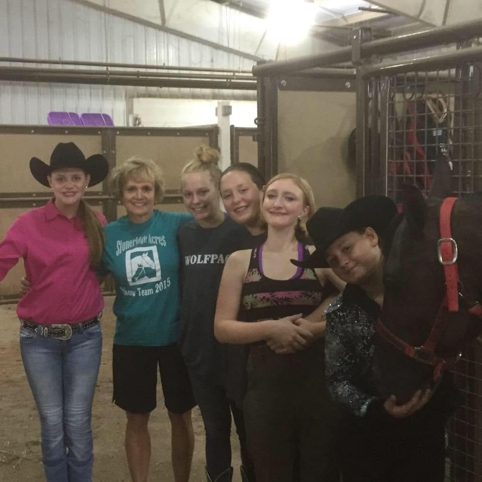 A group of young riders and an instructor pose in the barn aisle.